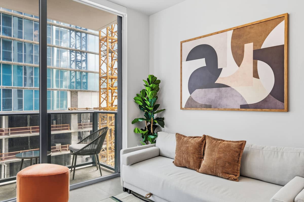 Un salon moderne avec un canapé, un grand tableau abstrait, une plante en pot et une vue sur un chantier de construction, visible à travers une porte vitrée.