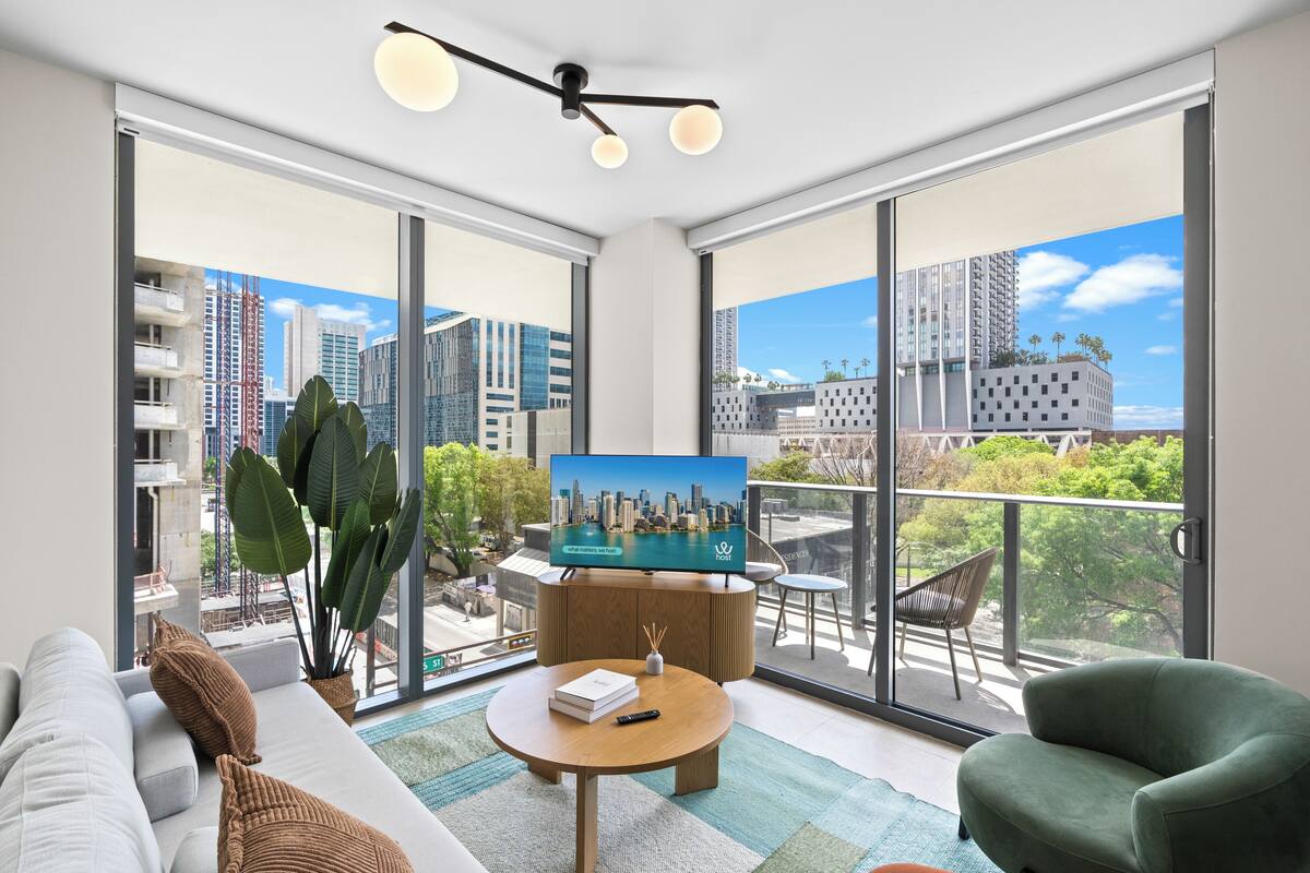 Un salon moderne avec une grande télévision, un canapé, un fauteuil vert et un balcon offrant une vue sur la ville.