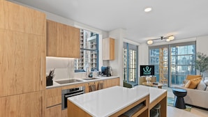 A modern kitchen with wooden cabinets, a white countertop, and a built-in oven. There is a dining area with a white table and chairs, and a sofa with a view of the city through large windows. - 11 Ne Worldcenter Experience - We Host (Miami)