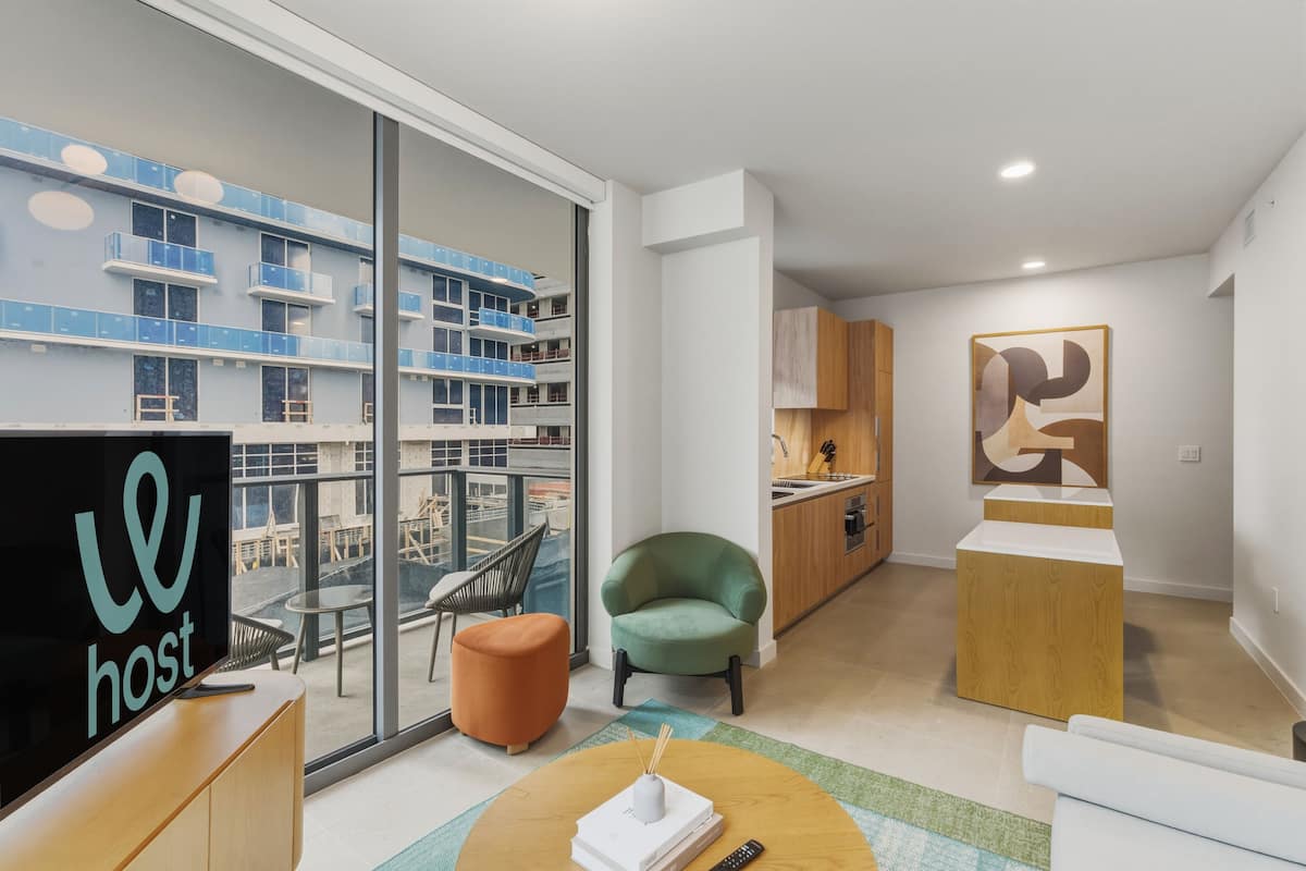 Un appartement moderne doté d’une grande porte-fenêtre coulissante donnant sur un balcon, d’un coin cuisine et d’un espace de vie comprenant un téléviseur, des sièges et un élément décoratif.