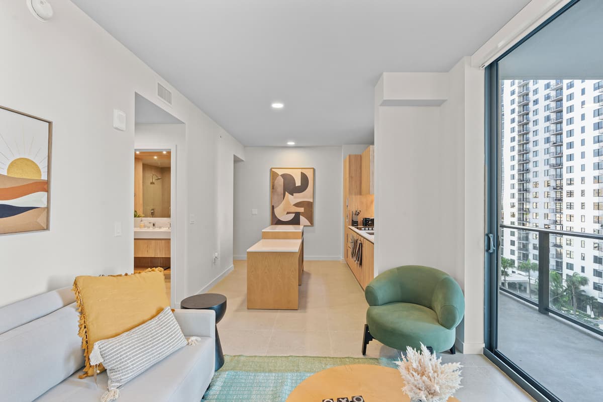Un salon moderne avec un canapé, un fauteuil vert, une table basse en bois et une vue sur un immeuble de grande hauteur, visible à travers une grande fenêtre.