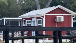 Un edificio rojo con puerta y ventanas blancas, un área cercada con un banco, y un árbol al fondo.