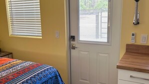 Un dormitorio con cama, una puerta, ventana con persianas, mesita de noche y lámpara de pared.