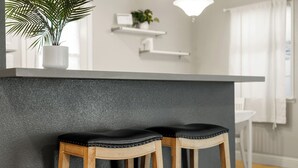 A kitchen area with a grey countertop, two wooden stools with black cushions, a potted plant, and white shelves with a small plant and a clock.