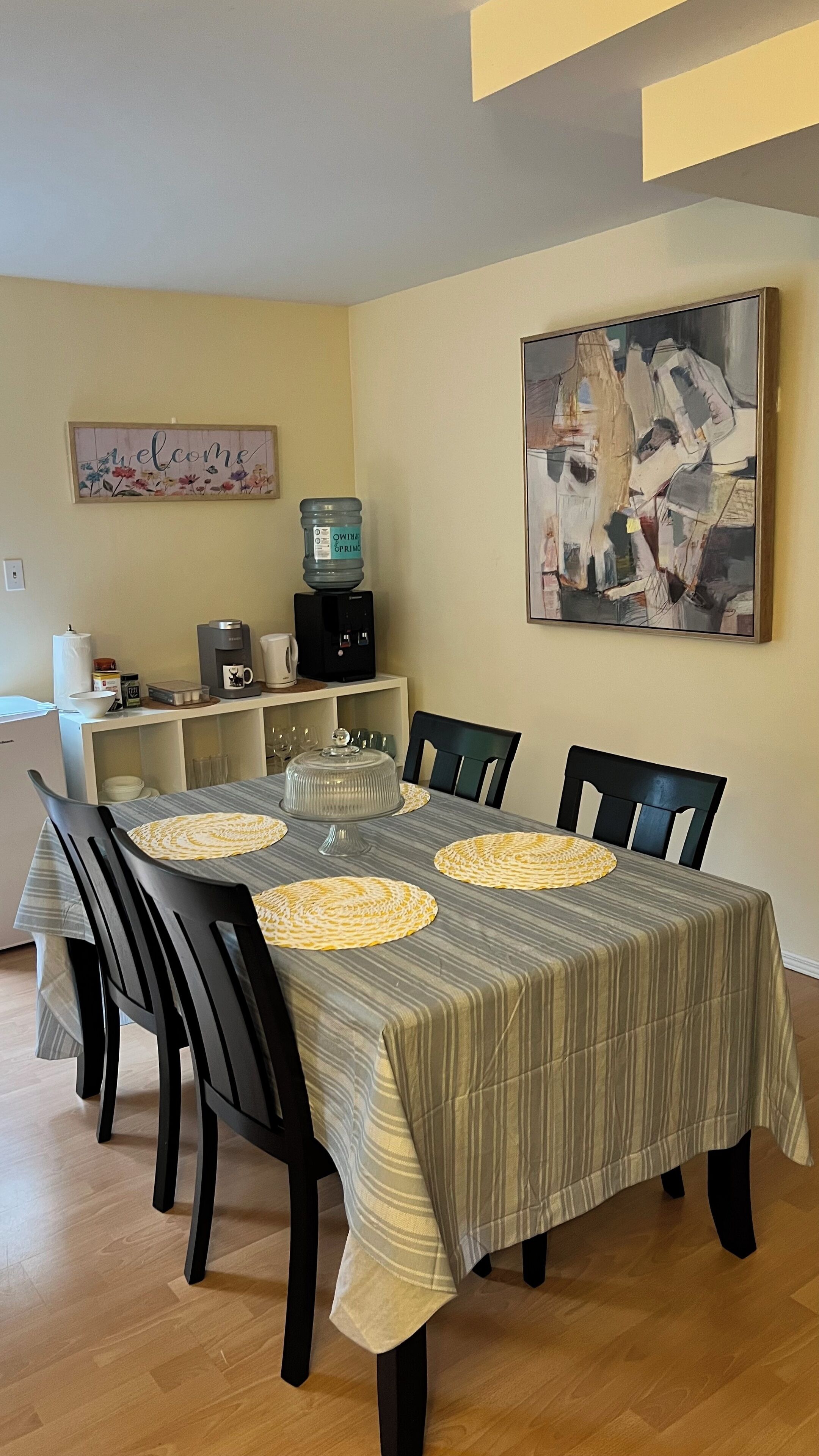 Un comedor con mantel a rayas, sillas negras, una estación de café y un letrero de bienvenida.