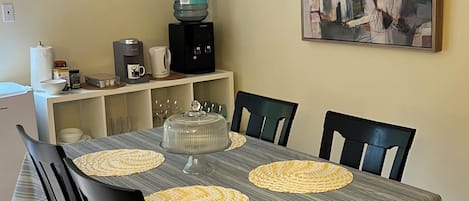 Un comedor con mantel a rayas, sillas negras, una estación de café y un letrero de bienvenida.