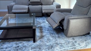 Un salon avec un canapé d’angle gris, une table basse en verre et un tapis à poils longs bleu.