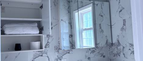 A bathroom with marble-patterned walls, a toilet, a sink with a mirror above it, and shelves with towels and toiletries.