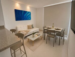 Een moderne woonkamer met een bank, salontafel, eettafel en stoelen. Aan de muur hangt een schilderij en rechts staat een televisie.