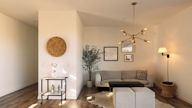 A modern living room with a wooden floor, a round woven wall art, a glass coffee table, a sofa with patterned cushions, a hanging light fixture, and a small side table with a lamp.