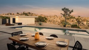 An outdoor dining area with a pool, desert landscape, and a small building.