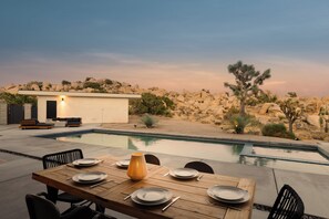 An outdoor dining area with a pool, desert landscape, and a small building.