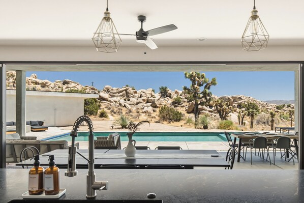 A modern kitchen with a view of a desert landscape, a pool, and outdoor seating.