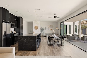 A modern kitchen with dark cabinetry, a marble island, and a dining area with a large table and chairs. - Mars Landing - Pool / Spa / Boulders / View / BBQ / Stargazing (Yucca Valley)