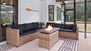 Een moderne buitenruimte met een wicker loungeset, een houten tafel met vuurkorf, en grote ramen met uitzicht op de natuur.