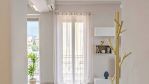 Ein Zimmer mit einem großen Fenster, das mit transparenten Vorhängen bedeckt ist, einer Topfpflanze, einer kleinen weißen Bank und einer dekorativen goldfarbenen Baumskulptur.
