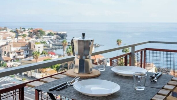 Ein Esstisch ist mit Tellern, Besteck und einer Kaffeekanne gedeckt, mit Blick auf eine Küstenstadt auf einem Balkon.