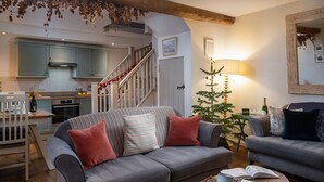 A cozy living room with a grey sofa, a small coffee table with books and a teapot, a Christmas tree, and a staircase leading to a kitchen.
