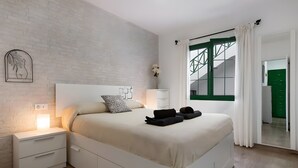 Un dormitorio moderno con pared de ladrillo blanco, cama con cabecera blanca, mesita de noche con cajones, mesita auxiliar con lámpara y vista a una puerta verde y una escalera.