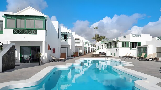 Zona de piscina con edificios blancos, cielo despejado y una palmera.