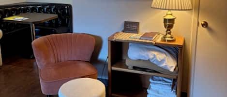 Una habitación con piso de madera, un sillón marrón, un pequeño reposapiés con estampado floral, una lámpara, un estante con ropa doblada y un espejo enmarcado sobre una ventana.