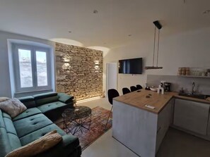 A modern living room with a green sofa, a stone accent wall, a dining area with a wooden table, and a kitchen with a TV and pendant lights. - Casa Arena 1 min walk to the Colosseum, city centre and harbour promenade 3 min (Pula)