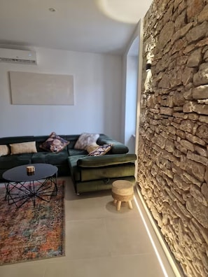 A modern living room with a green sofa, a round coffee table, a small stool, a patterned rug, and a stone wall. - Casa Arena 1 min walk to the Colosseum, city centre and harbour promenade 3 min (Pula)