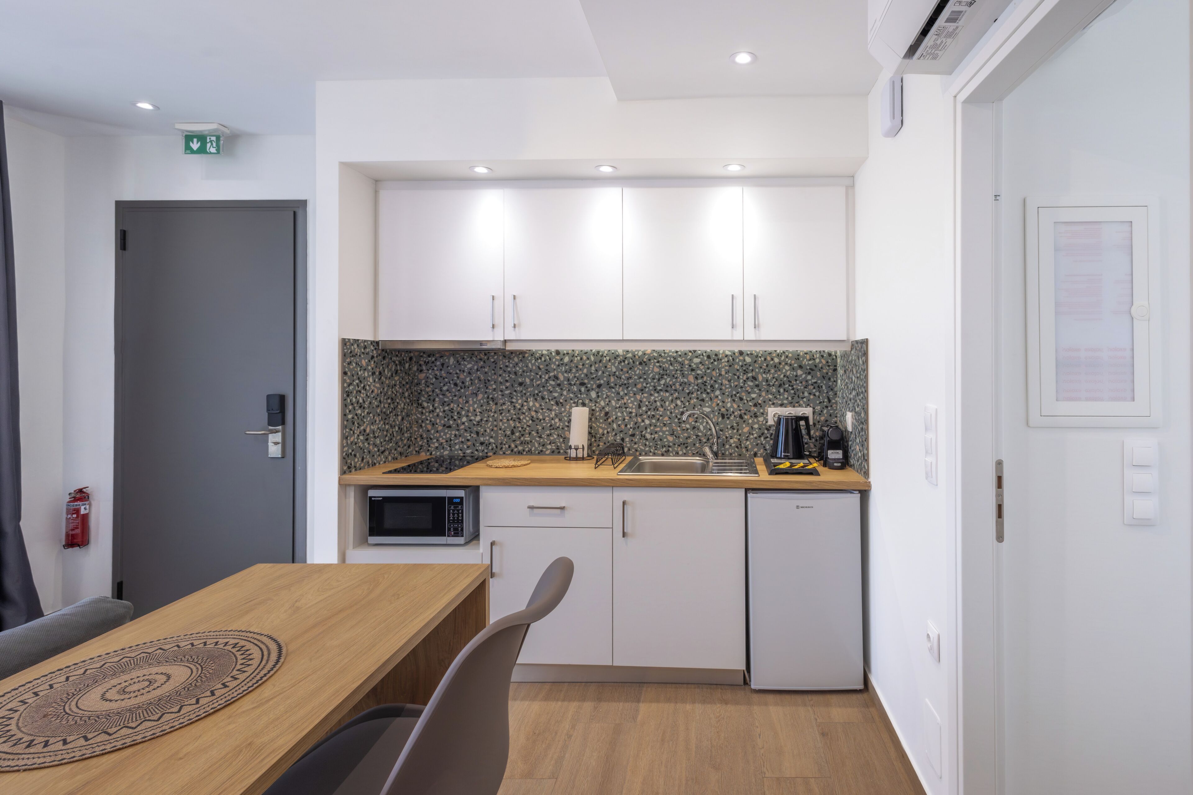 A modern kitchen with white cabinets, a granite backsplash, a microwave, a coffee maker, and a small refrigerator.
