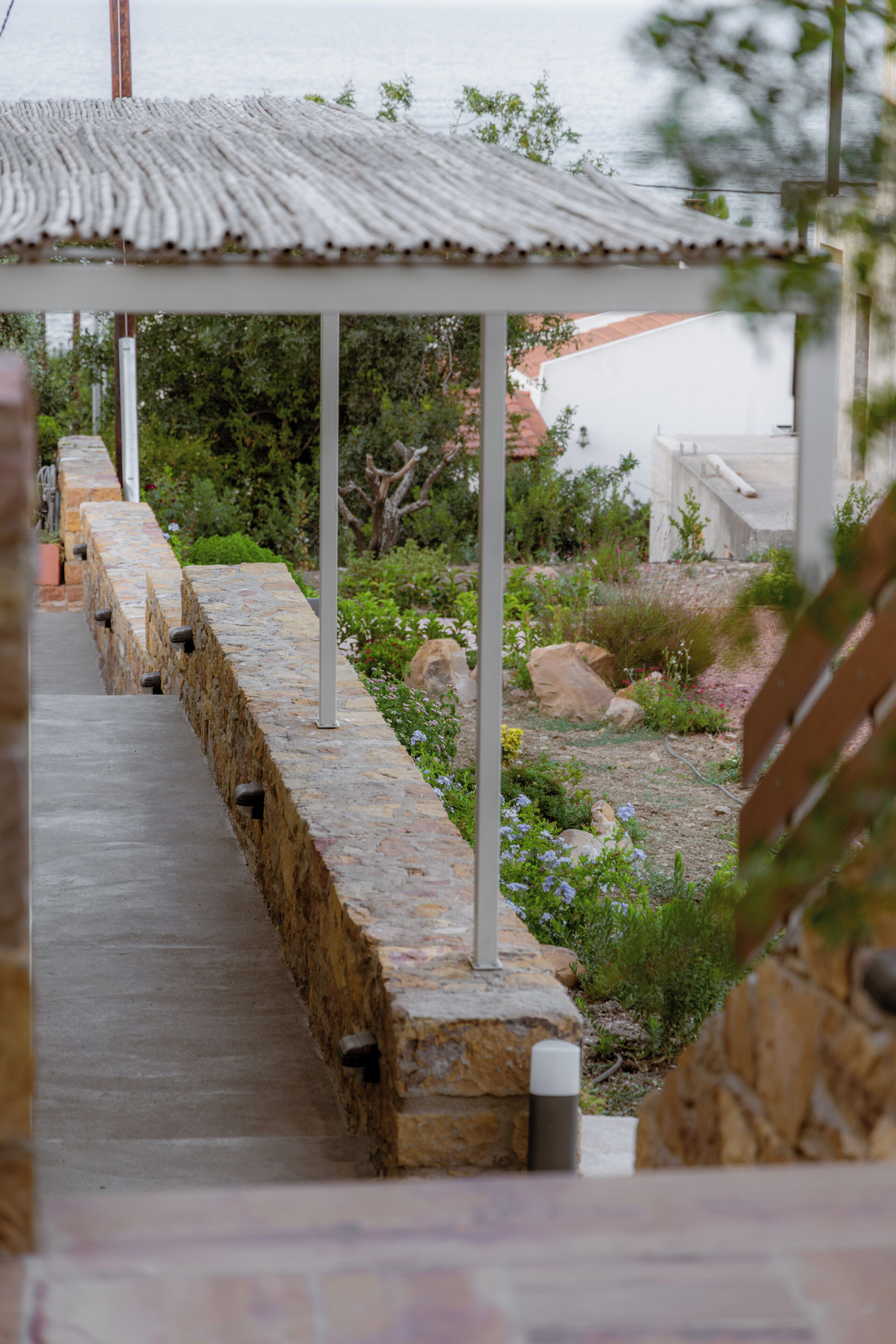 A stone pathway leading to a covered area with a metal roof, surrounded by greenery and a body of water.