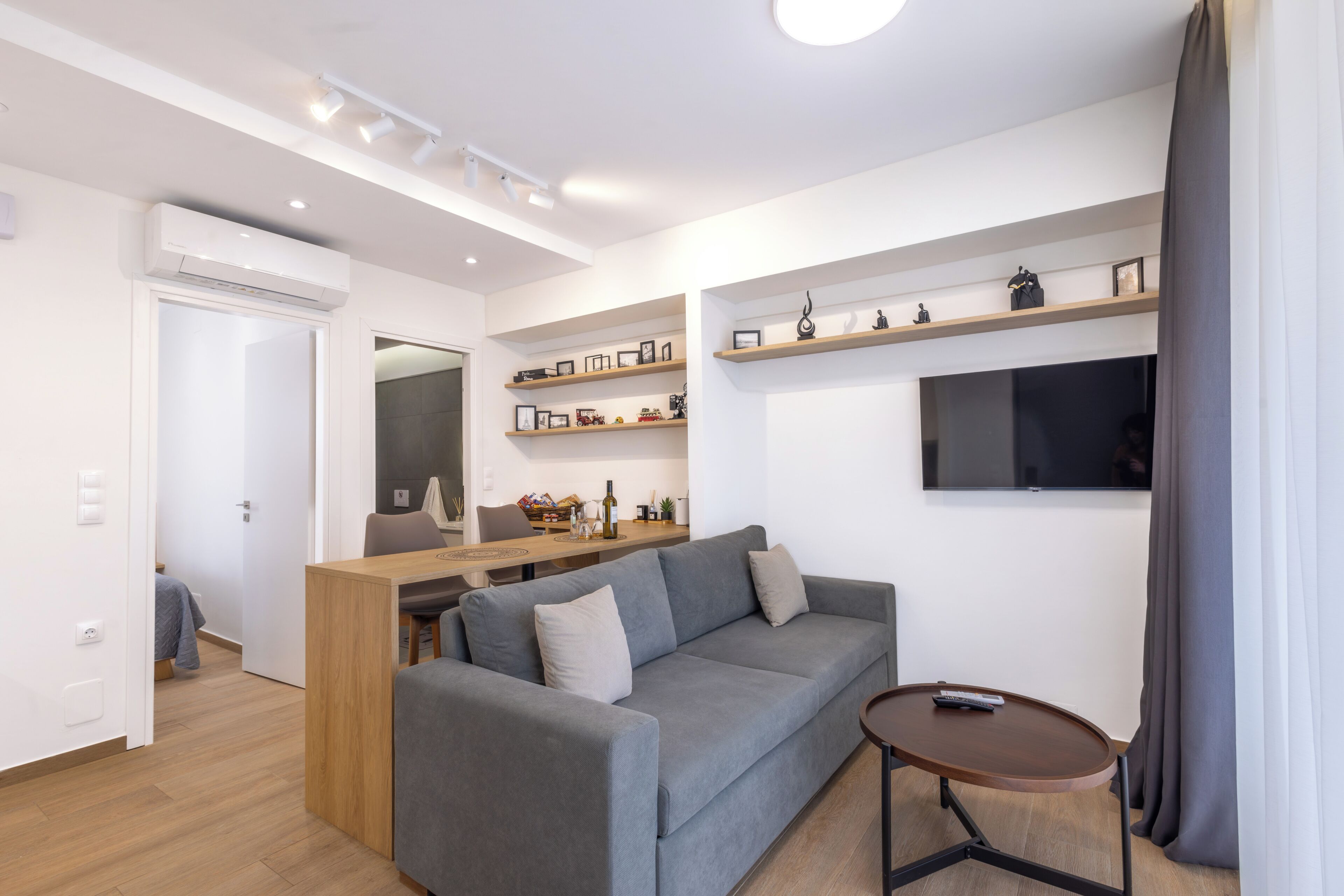 Una sala de estar moderna con un sofá gris, una mesita redonda, un televisor montado en la pared y un escritorio de madera con estanterías arriba.