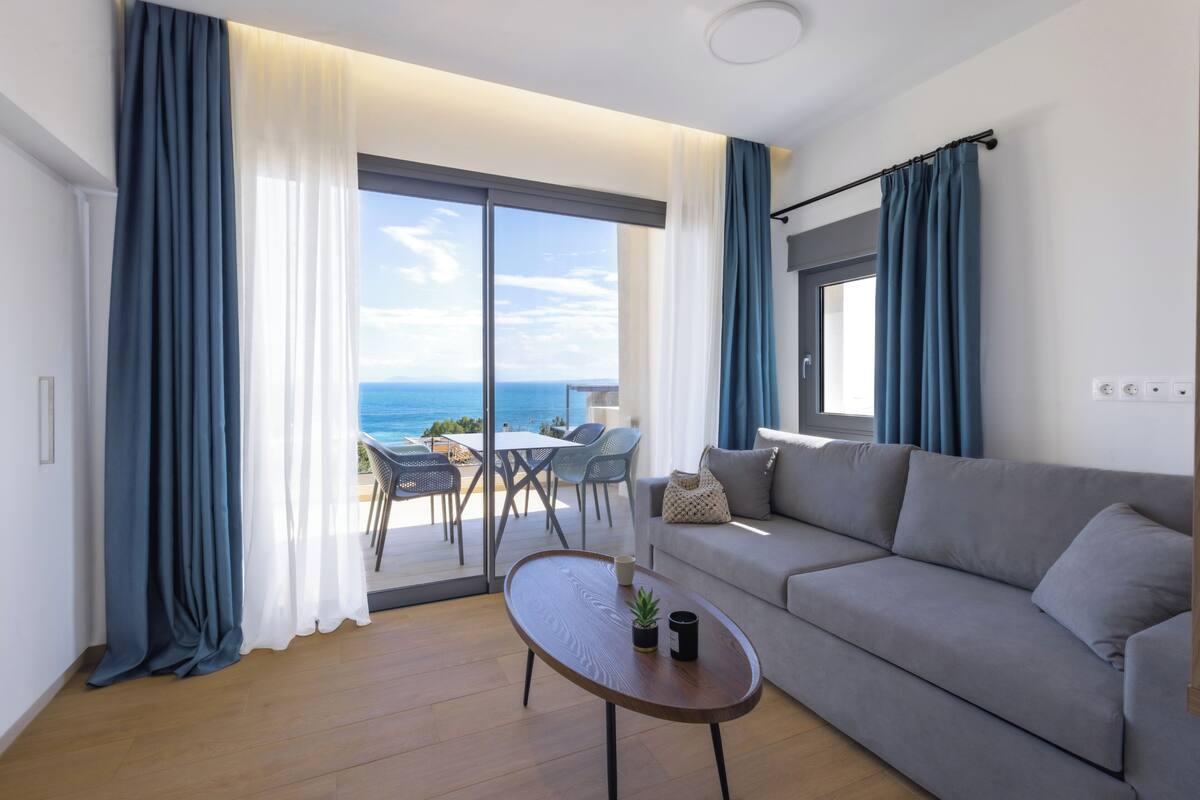 A modern living room with a grey sofa, a wooden coffee table, and a view of the ocean through large windows.