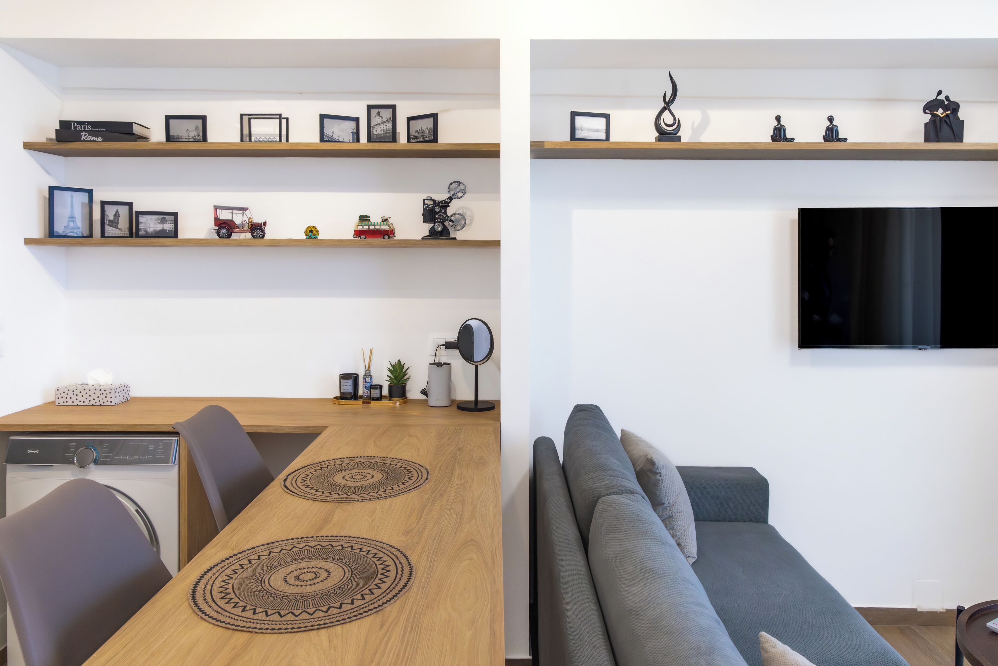 Un espai de vida modern amb zona de menjador, sofà, televisió penjada a la paret i prestatges amb diversos objectes.