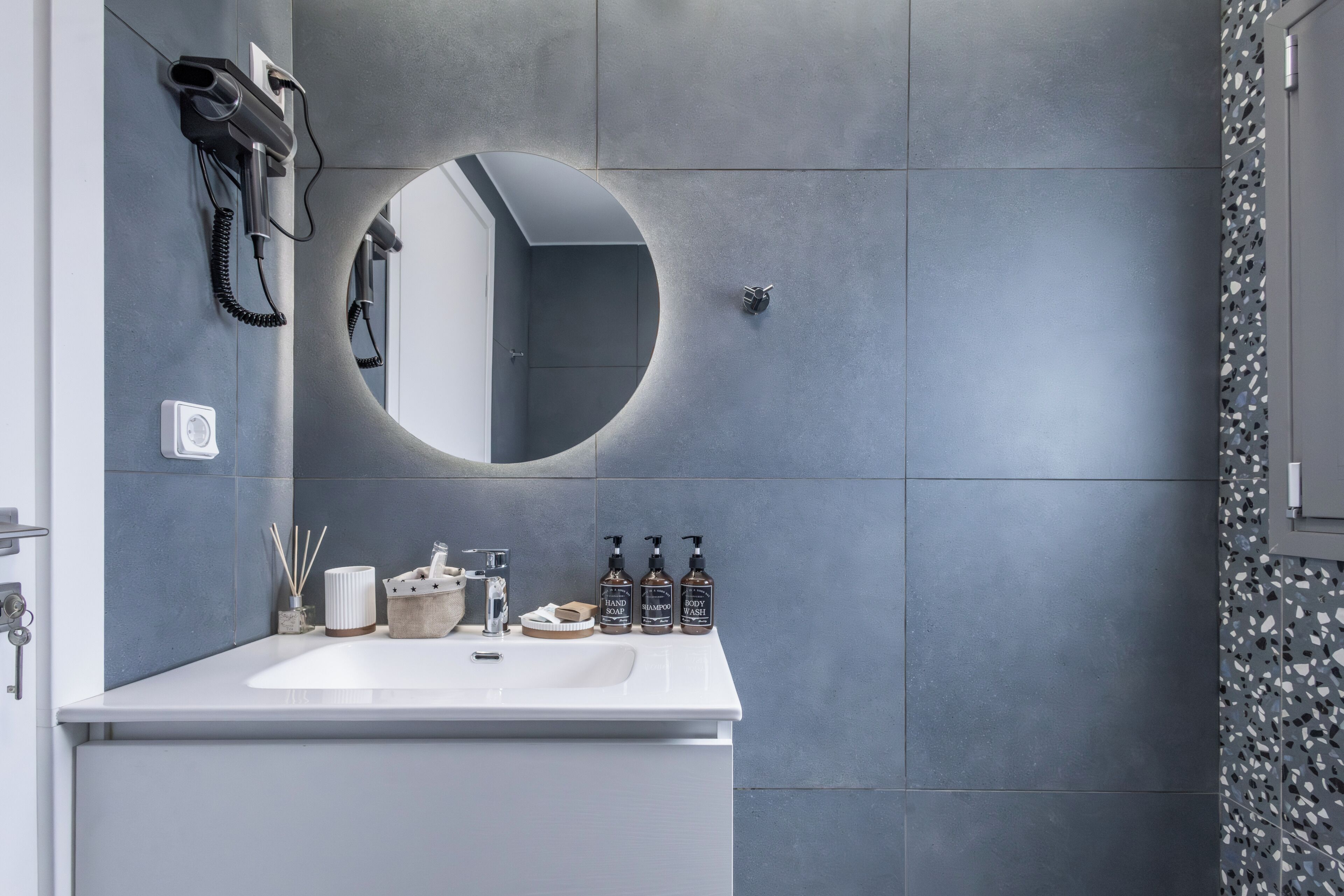 Un baño moderno con un lavamanos blanco, un espejo redondo y artículos de aseo en la encimera.