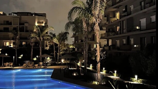 A nighttime view of a hotel pool area with multiple floors of buildings, palm trees, and lounge chairs. - Stylish 3BR Serviced Aprt Pool View Sodic Villette (Cairo Governorate)