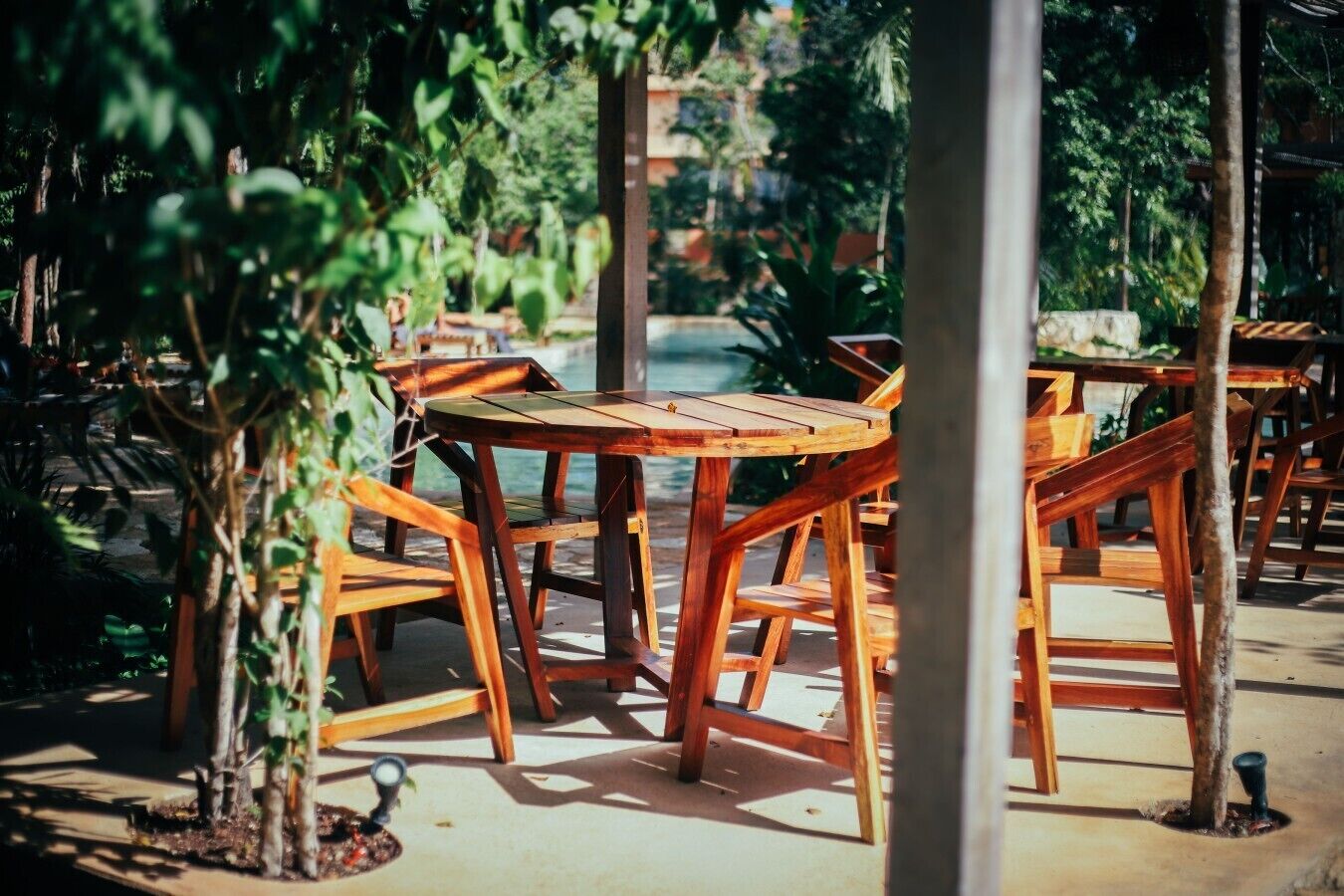 Outdoor wooden tables and chairs arranged in a shaded area with a view of a body of water.