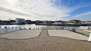 A waterfront promenade with a paved walkway, a wooden fence, and a view of buildings across the water. - Boater & Beach Lover’s Retreat (Manahawkin)