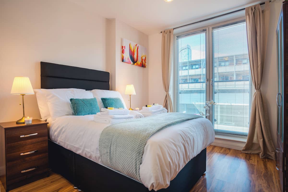 A modern hotel room with a large bed, two bedside tables with lamps, a nightstand, a window with blinds, and a painting on the wall.