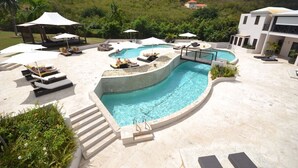 A pool area with lounge chairs, umbrellas, and a building in the background. - Villa Kokomo · Breathtaking views 4BR Luxury Villa in Sugar Ridge w Private Pool (Jolly Harbour)
