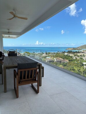 A balcony with a wooden table and chairs, a ceiling fan, and a view of the ocean and a coastal town.