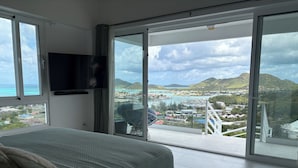 A bedroom with a bed, a TV, and a view of a coastal town through large windows. - Villa Kokomo · Breathtaking views 4BR Luxury Villa in Sugar Ridge w Private Pool (Jolly Harbour)