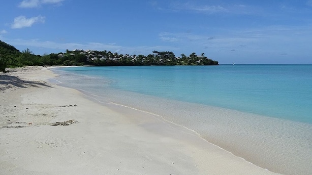 A beach with clear turquoise water, a sandy shore, and a small island with buildings in the background. - Villa Kokomo · Breathtaking views 4BR Luxury Villa in Sugar Ridge w Private Pool (Jolly Harbour)
