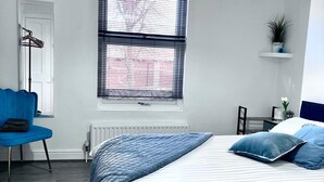Ein Schlafzimmer mit einem Bett, einem blauen Sessel, einem Heizkörper, einem Fenster mit Jalousien und einer Pendelleuchte.