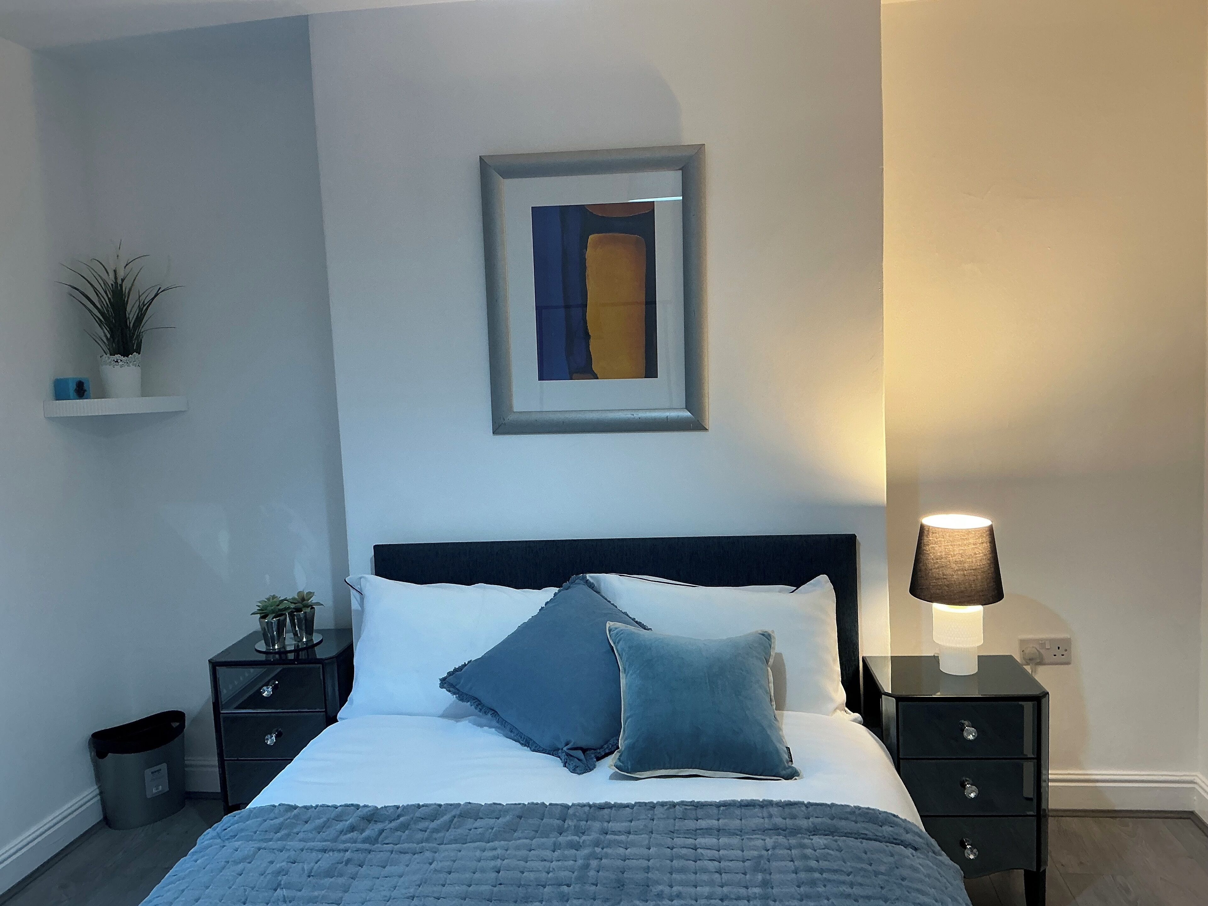 Un dormitorio con cama, una mesita de noche con lámpara, un estante en la pared con plantas y un cuadro enmarcado.