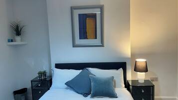 Un dormitorio con cama, una mesita de noche con lámpara, un estante en la pared con plantas y un cuadro enmarcado.