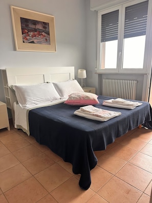 A bedroom with a bed, pillows, a nightstand, a lamp, a window with blinds, and a radiator.