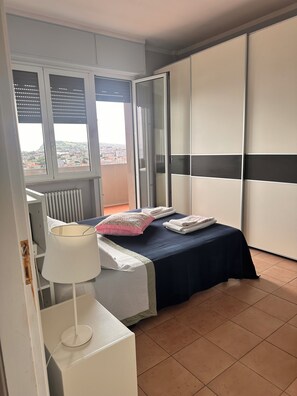 A bedroom with a bed, a nightstand, a lamp, a radiator, and a view of the city through a window. - Waterfront apartment with breathtaking views in a very central location. (Pesaro)