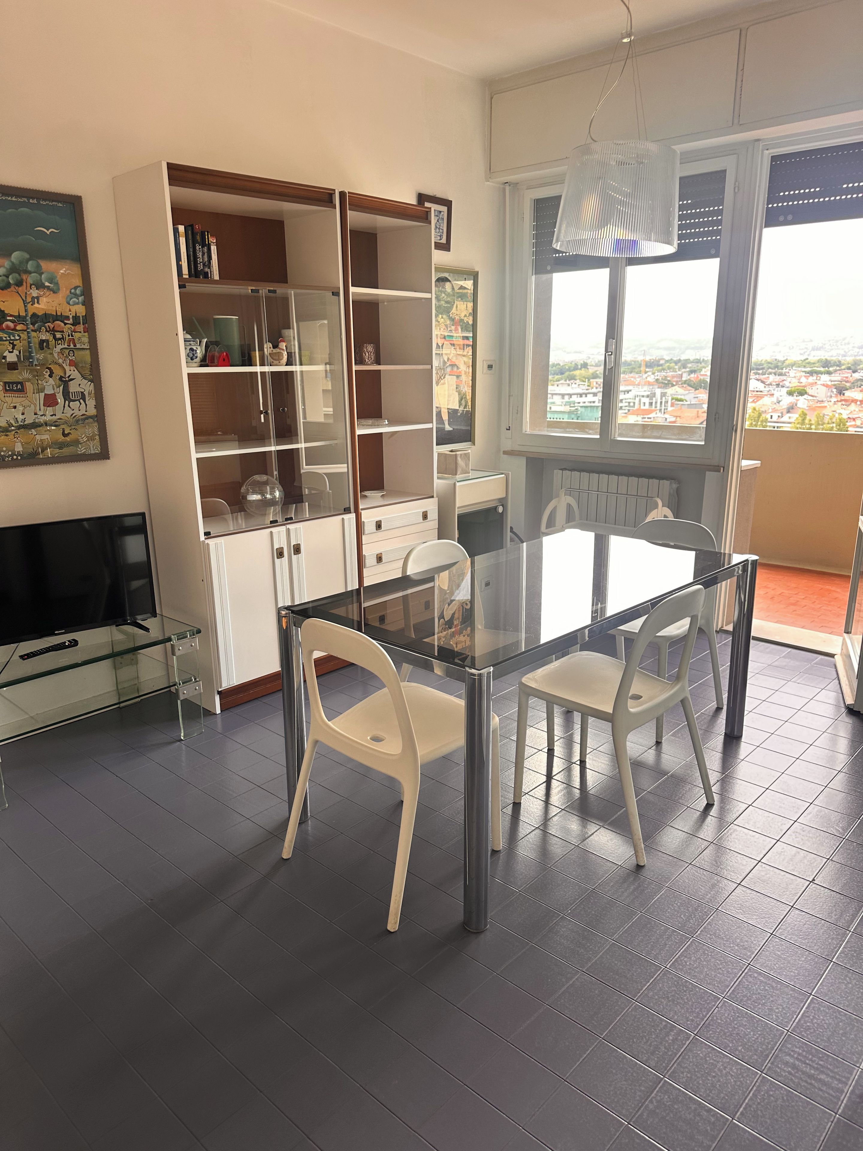 A modern living room with a glass dining table, white chairs, a TV, and a large window with a city view.