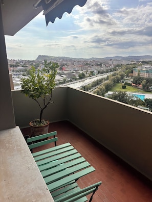 A balcony with a potted tree, a green bench, and a view of a cityscape with buildings and a river. - Waterfront apartment with breathtaking views in a very central location. (Pesaro)