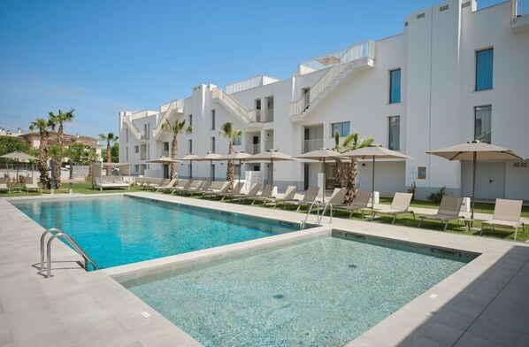 A swimming pool with lounge chairs and umbrellas, surrounded by white buildings with balconies.