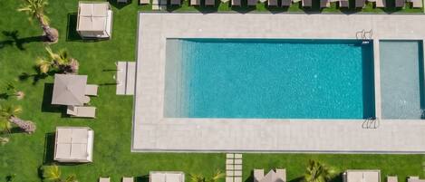 Vista aerea di una piscina rettangolare circondata da lettini e ombrelloni, con un vialetto lastricato e palme.